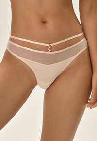 Beżowe stringi z przezroczystymi panelami z siateczki, z podwójnymi dekoracyjnymi paskami i okrąłym akcentem na środku. Gładkie wykończenie z minimalnym pokryciem.