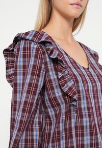 Bordeaux geruite blouse met volantren, een V-halslijn en een mix van fijne blauwe en witte lijnen in het patroon. Zachte textuur.