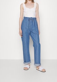 Pantaloni di jeans blu, a vita alta, a gamba larga con vita regolabile. Abbinati a una canotta bianca a costine e sandali bianchi slip-on.
