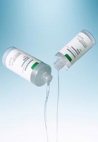 Dwie przezroczyste butelki z plastiku, oznaczone napisem "Peeling eksfoliujący", wylewają przezroczysty płyn na jasnoniebieskim tle, prezentując elegancki design i minimalistyczny tekst.