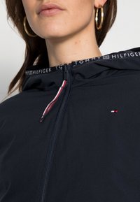 Giacca nera con cappuccio e zip frontale, dotata di nastro al collo con logo e accenti rosso, bianco e blu. Texture liscia, design aderente.
