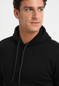 Zwarte hoodie van zacht materiaal, voorzien van een trekkoord met witte accenten en een buidelzak. Klassieke pasvorm met een geribbelde halslijn.