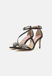 Black stiletto heels with suede straps, featuring a prominent embellished detail at the front and a delicate ankle strap for support.