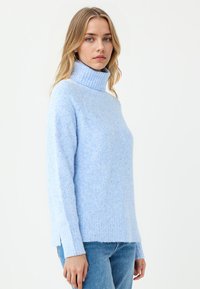 Hellblaues Sweatshirt aus weichem, strukturiertem Material mit hohem geripptem Kragen und seitlichen Schlitzen; kombiniert mit blauen Jeans.