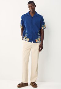 Chemise bleu à manches courtes avec broderies tropicales, pantalon beige et sandales marron à enfiler. La chemise présente un col à revers et une fermeture à boutons sur le devant.