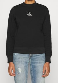 Schwarzer Pullover mit Rundhalsausschnitt, gerippten Bündchen und Saum, mit einem silbernen "cK"-Logo auf der Brust. Kombiniert mit hellblauen, verwaschenen Jeans.