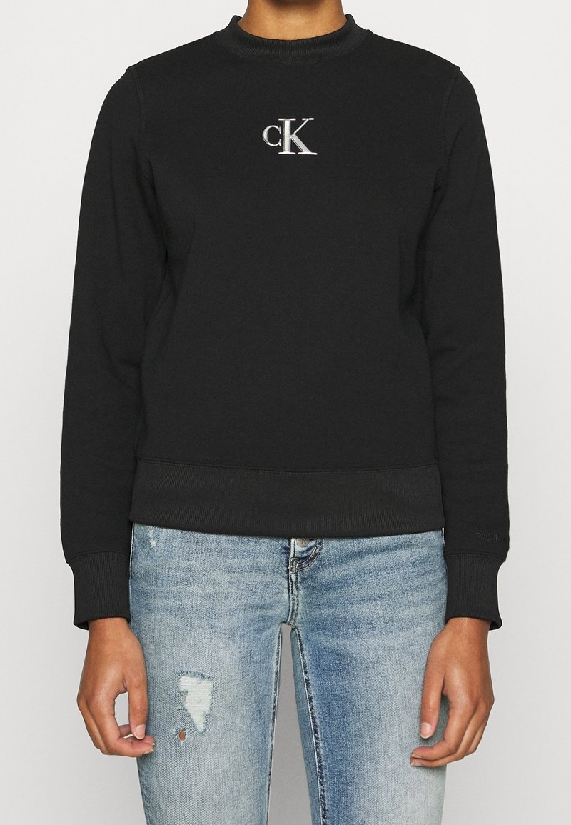 Schwarzer Pullover mit Rundhalsausschnitt, gerippten Bündchen und Saum, mit einem silbernen "cK"-Logo auf der Brust. Kombiniert mit hellblauen, verwaschenen Jeans.