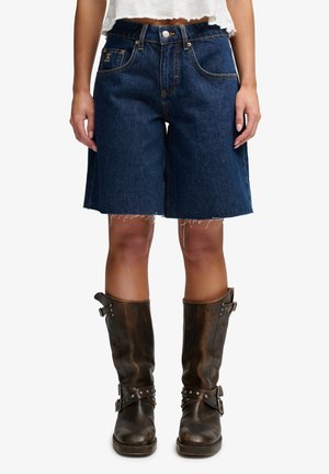 Person iført mørkeblå cowboyshorts til knæet med rå kant og brune læder midt på læggen støvler med spænder og nitter.