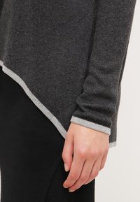 Main reposant sur le côté, vêtue d'un pull gris foncé avec une bordure gris clair et d'un pantalon noir avec un détail de couture discret.