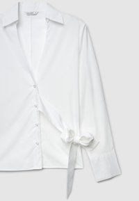 White shirt with a tied bow detail at the waist, button front, and long sleeves. Made from smooth fabric with a classic collar and cuffs.