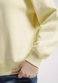 Żółta bluza z fakturowanym wykończeniem, ściągaczami przy mankietach i u dołu. Małe logo widoczne w pobliżu dolnej krawędzi. Noszona na dżinsach.