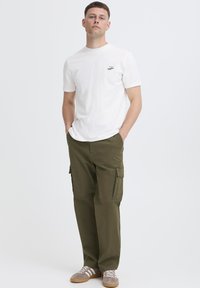 Valkoinen lyhythihainen t-paita, jossa pieni logo, yhdistetty oliivinvihreisiin cargo-housuihin, joissa on sivutaskut. Asun viimeistelevät harmaat ja valkoiset tennarit.