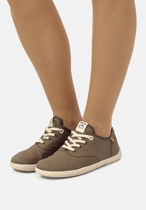 Zapatillas de lona marrones con suelas y cordones blancos, llevadas en los pies sobre un fondo claro y liso.