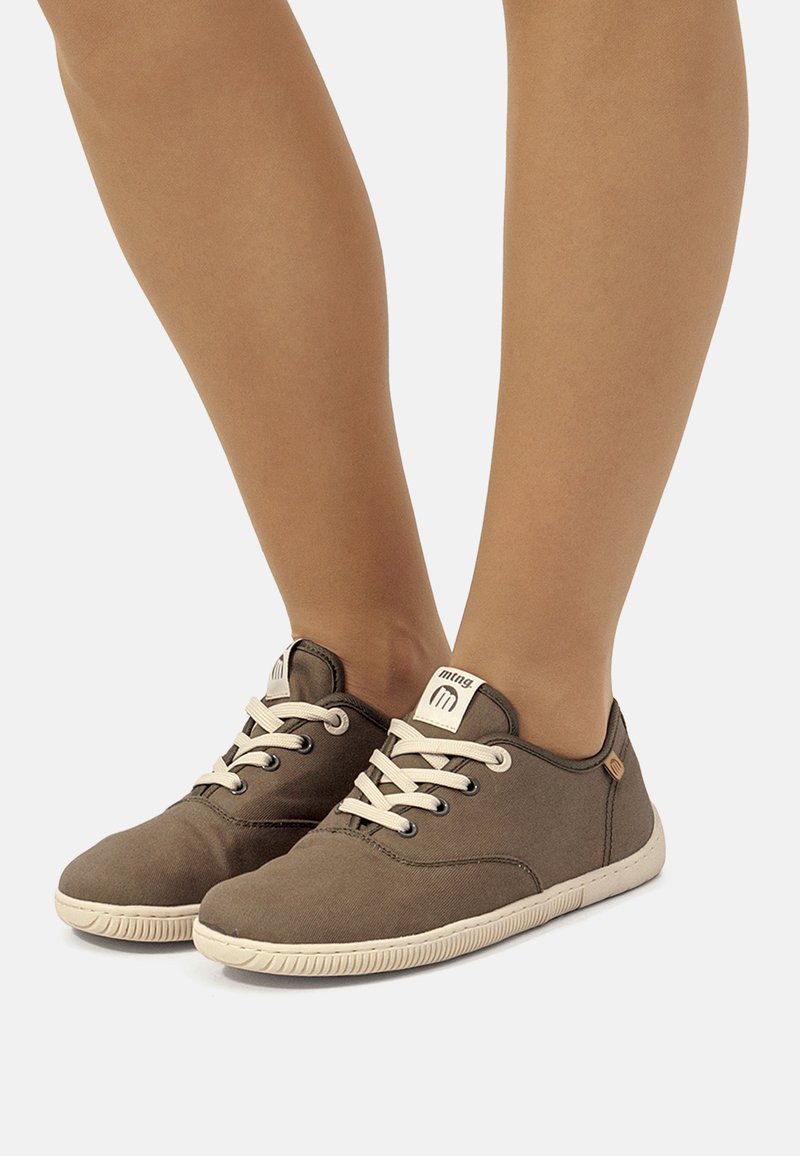 Zapatillas de lona marrones con suelas y cordones blancos, llevadas en los pies sobre un fondo claro y liso.