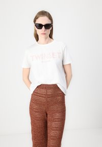 Valge puuvillane T-särk, millel on roosade tähtedega "TWINSET MILANO"; koos rustikarva heegeldatud pükstega, millel on tekstuuriline mustri. Mustad kassisilma prillid.
