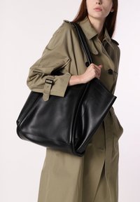 Zwarte leren tote bag met een gestructureerd ontwerp, met een gladde textuur en contrasterende stiksels, vastgehouden door een persoon in een beige jas.