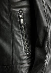 Black leather jacket featuring a zipped pocket with silver hardware and textured surface. Detail stitching visible near the pocket.