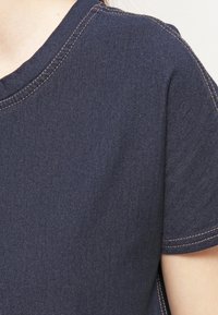 Detailní záběr osoby v tmavě modrém tričku s krátkým rukávem, na kterém jsou viditelné béžové stehy podél krku, ramene a švů rukávu.
