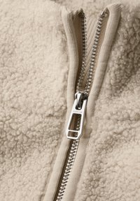 Beige fleecejack met een gestructureerd oppervlak, voorzien van een zilveren metalen rits met een rechthoekige trekker.