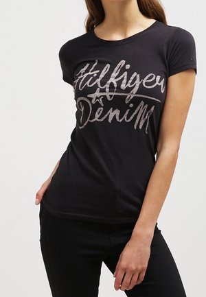 Kobieta nosząca dopasowany czarny t-shirt z napisem „Hilfiger Denim” dużymi, swobodnymi literami na piersi, w połączeniu z czarnymi spodniami.