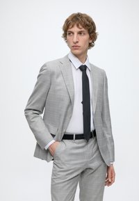 Jeune homme portant un costume à carreaux gris clair, chemise blanche et cravate noire, regardant sur le côté, une main dans la poche, sur un fond uni.