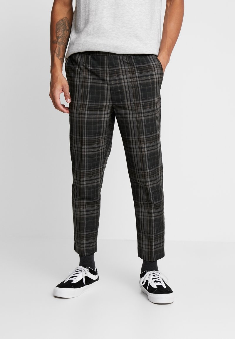 Uomo che indossa pantaloni a scacchi neri e grigi con calzini neri e sneaker bianco e nere, in piedi con le mani nelle tasche.