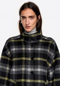 Abrigo de lana a cuadros con un alto cuello y botones negros. Los colores dominantes incluyen negro, gris, y toques de amarillo y blanco.