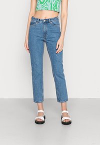 Jeans de ganga azul-claro com embellishments semelhantes a pérolas, cintura alta, corte reto e bainha desfeita. Combinados com sandálias brancas num piso neutro.