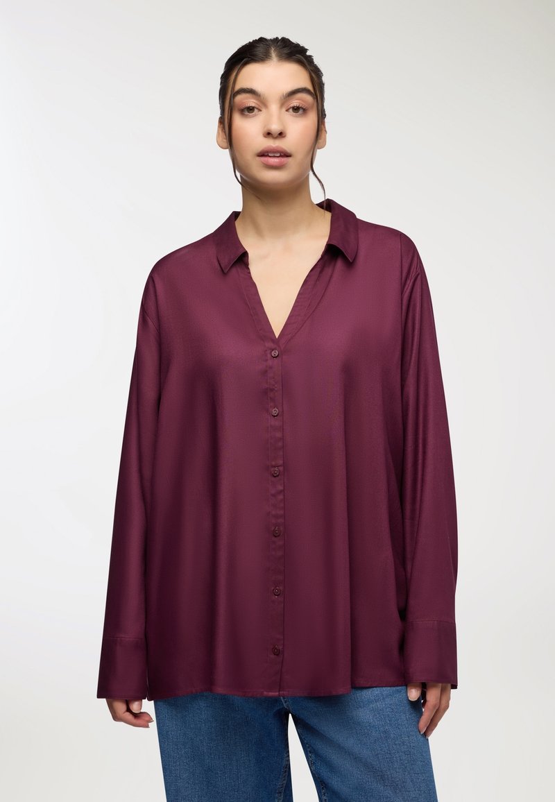 Blusa oversize color marrone realizzata in tessuto liscio, dotata di scollatura a V, maniche lunghe e chiusura frontale con bottoni, abbinata a jeans azzurri chiari.