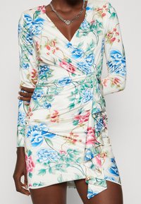 Robe portefeuille florale avec un fond crème, motifs de fleurs bleues et roses, manches longues et un ourlet à volants. Le matériau semble lisse.