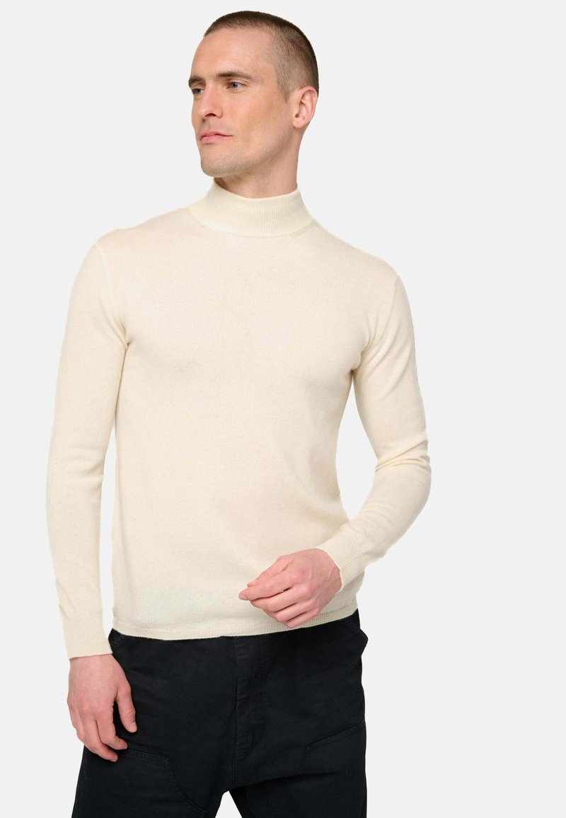 Homme portant un pull col roulé crème ajusté et un pantalon noir, posant avec une main près de la taille contre un fond clair.