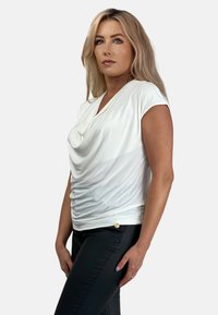 Biała bluzka z krótkim rękawem, z marszczonym wykończeniem z przodu i złotym guzikiem na dole, w połączeniu z czarnymi spodniami.