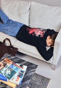 Ragazzo adolescente sdraiato a faccia in giù su un divano beige, indossando jeans e un felpa nera con grafica rossa, accanto a un tavolino in marmo con libri impilati.