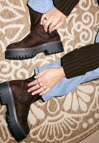 Stivaletti ankle in suede marrone con suola in gomma nera spessa. Il design presenta una cerniera laterale e una suola testurizzata. Sono visibili mani con anelli.