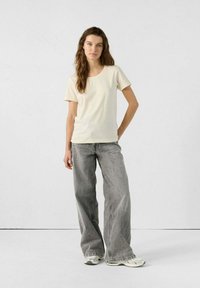 Camiseta de manga corta en beige claro combinada con unos pantalones vaqueros grises de tiro alto y pierna ancha. El conjunto presenta líneas simples y limpias, así como texturas suaves.
