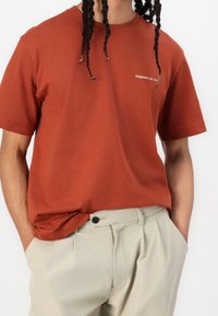 Camiseta de algodón color óxido, con mangas cortas y escote redondo. Presenta una pequeña impresión de texto blanco en el pecho. Se lleva con pantalones beiges.