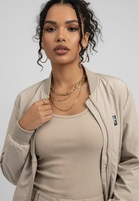 Beige Bomberjacke mit gerippten Bündchen, getragen über einem beigen, gerippten Tanktop. Sie verfügt über geschichtete goldene Kettenhalsketten und ein kleines Logodetail.