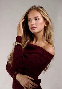 Maglione borgogna off-shoulder con texture a coste, orologio dorato e orecchini a forma di cuore. Lunghe, lisce ciocche di capelli cadono delicatamente sulle spalle.