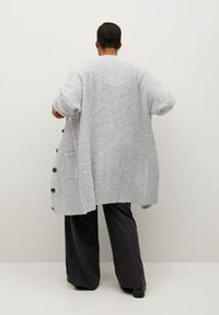 Grauer, strukturierter, übergroßer Pullover mit seitlichen Knöpfen und Taschen, kombiniert mit weiteren schwarzen Hosen. Rückansicht vor einem neutralen Hintergrund.