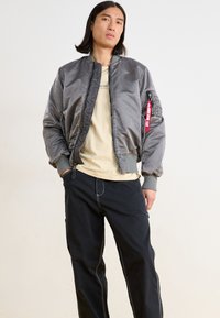 Blouson gris en satin, avec des poignets côtelés, un pantalon noir avec des coutures blanches et un T-shirt beige en dessous. Détail de l'étiquette rouge.