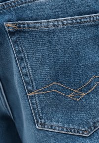 Πίσω τσέπη μπλε τζιν με αντίθετη πορτοκαλί ραφή και διακοσμητική λεπτομέρεια με ζιγκ-ζαγκ ραφή.