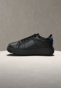 Μαύρο αθλητικό παπούτσι χαμηλού προφίλ με διάτρητες πλευρές και το κείμενο "LOVE MOSCHINO" στην εξωτερική πλευρά, τοποθετημένο πάνω σε φωτεινή πέτρινη επιφάνεια.