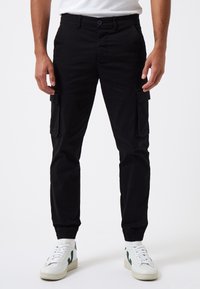 Pantalones cargo negros con puños elásticos, que cuentan con bolsillos laterales y un corte relajado. Combinados con zapatillas blancas que presentan detalles en verde.