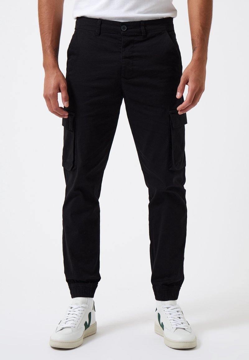 Pantalones cargo negros con puños elásticos, que cuentan con bolsillos laterales y un corte relajado. Combinados con zapatillas blancas que presentan detalles en verde.