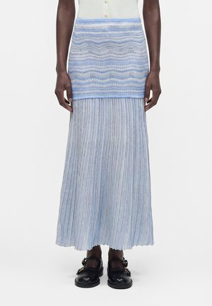 Μοντέλο φοράει μια πλισέ μάξι φούστα με μπλε και άσπρες ρίγες, με υφή και κοντύτερο μπλε επικάλυμμα και μαύρα παπούτσια με αγκράφα.