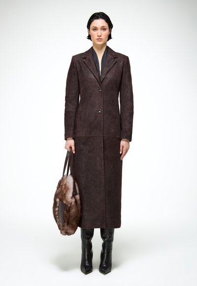 Femme portant un long manteau en cuir marron foncé, des bottes noires, tenant un grand sac à main en fourrure marron, debout devant un fond blanc uni.