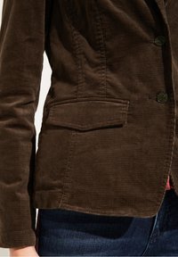 Ruskea samettinahkatakki, jossa on yksittäinen nappisuljenta, rintatasku ja räätälöity istuvuus jäsennellyillä harteilla.