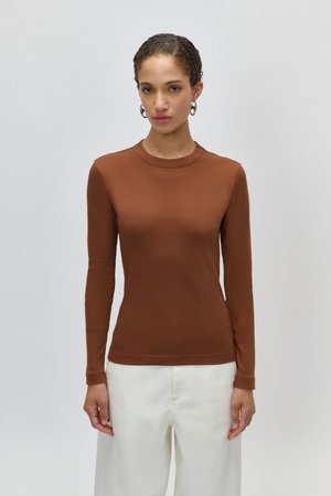 Donna con capelli ricci che indossa orecchini a cerchio, una maglia marrone aderente a maniche lunghe e pantaloni bianchi a vita alta, in piedi davanti a uno sfondo semplice.