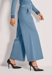 Hellblaue, weit geschnittene Hose mit flachem Bund und seitlichen Taschen, kombiniert mit schwarzen High Heels, die einen Riemen über den Fuß haben.