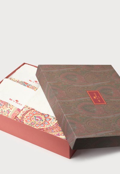 Boîte décorative partiellement ouverte avec un couvercle à motifs laissant voir des morceaux de tissu blanc pliés avec des bordures florales et cachemire colorées.